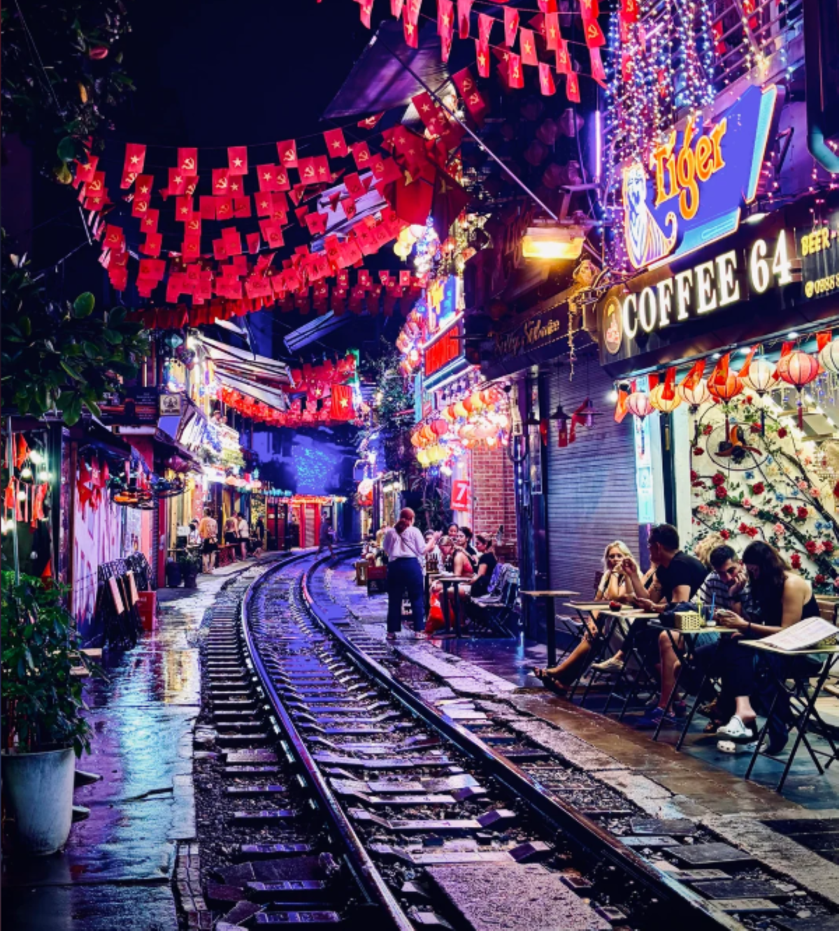 A lively street at night with train tracks running through outdoor cafes, illuminated by colorful lanterns and neon signs, and red flags hanging overhead. People are dining and walking along the track.
