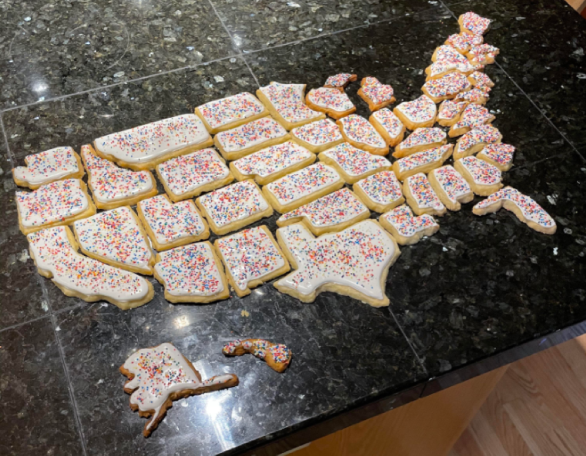 Cookies shaped like U.S. states, decorated with white icing and multicolored sprinkles, are arranged on a dark countertop to form a map of the United States.