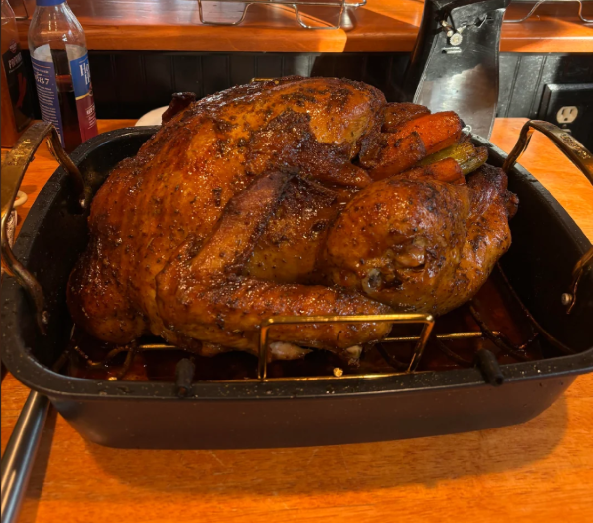 A golden-brown roasted turkey rests in a black roasting pan on a wooden countertop, with carrots and herbs visible inside the cavity.
