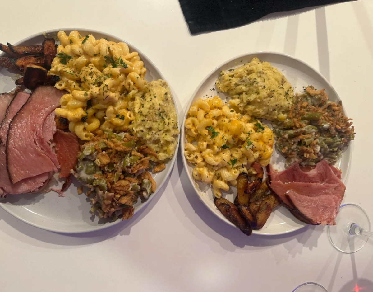 Two plates of food with macaroni and cheese, mashed potatoes, green bean casserole, roasted plantains, and slices of ham, set on a white table.