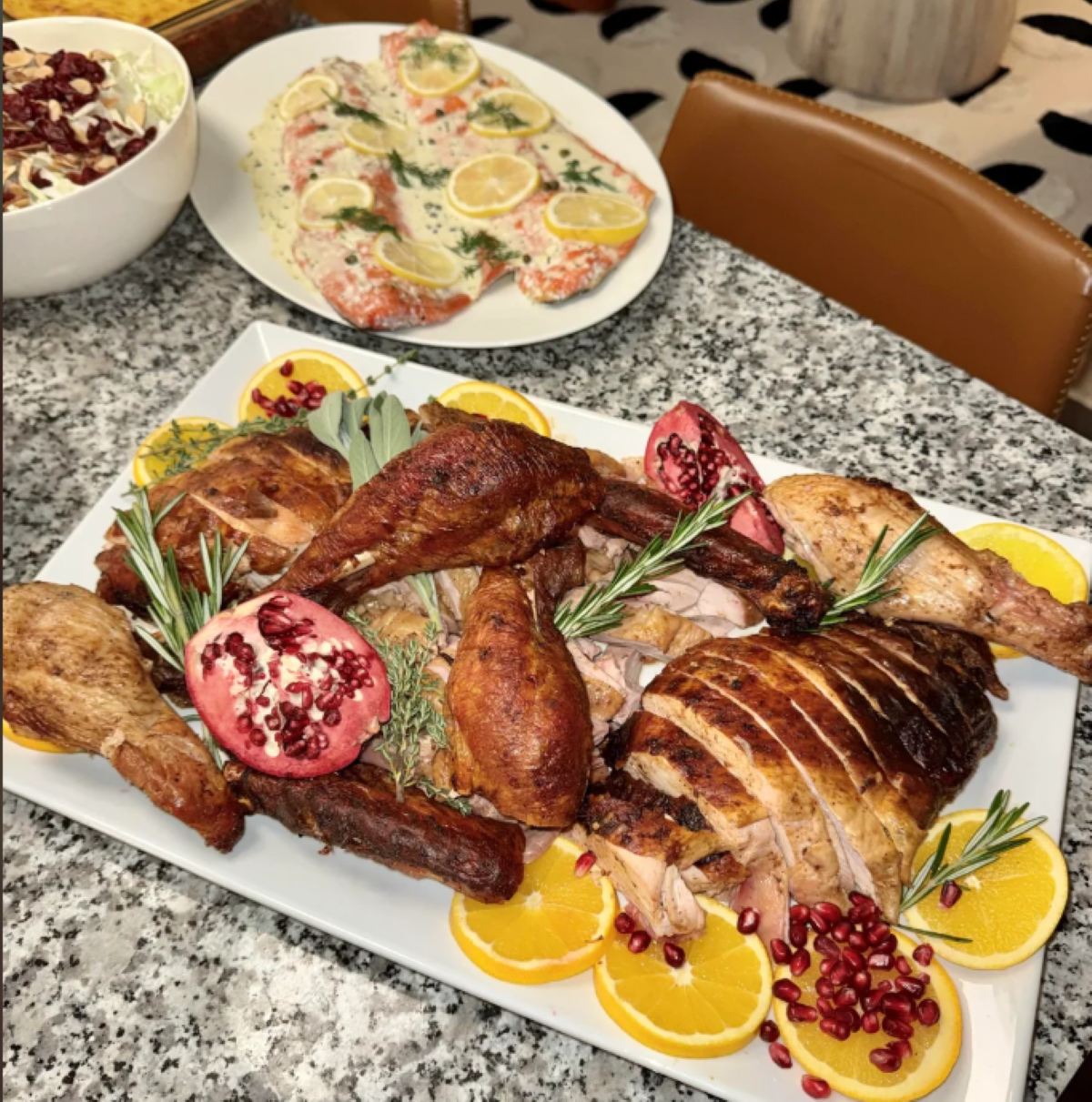 A platter of roasted turkey garnished with orange slices, rosemary, and pomegranate, with a side dish of salmon with lemon slices and a bowl of salad in the background on a marble table.