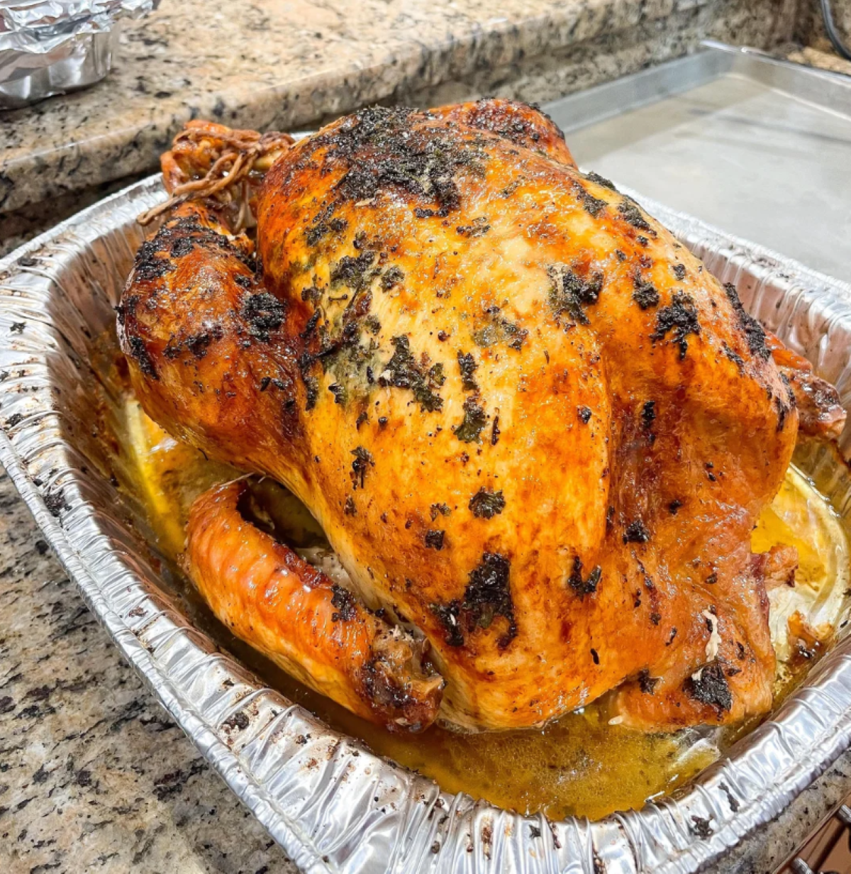 A whole roasted turkey with crispy, golden brown skin sits in a foil tray on a granite countertop. Herbs and juices cover the top, and the turkey is ready to be served.