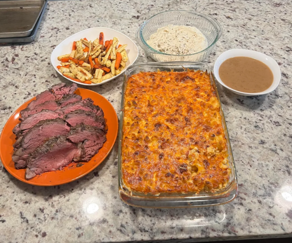 Sliced roast beef, roasted mixed vegetables, a baked cheesy casserole, a bowl of white rice, and a bowl of brown gravy are arranged on a granite countertop.