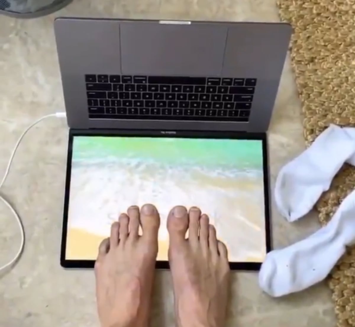 Bare feet rest on a laptop screen displaying a beach scene, simulating standing at the shore. A pair of white socks lies nearby on the floor next to the laptop.
