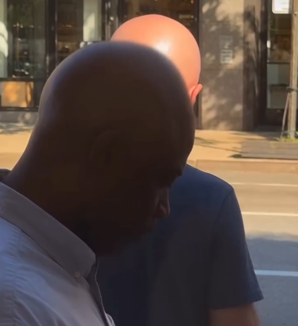 Two bald men stand on a sidewalk, one facing forward in shadow and the other with his back to the camera in sunlight. A city street and building windows are visible in the background.
