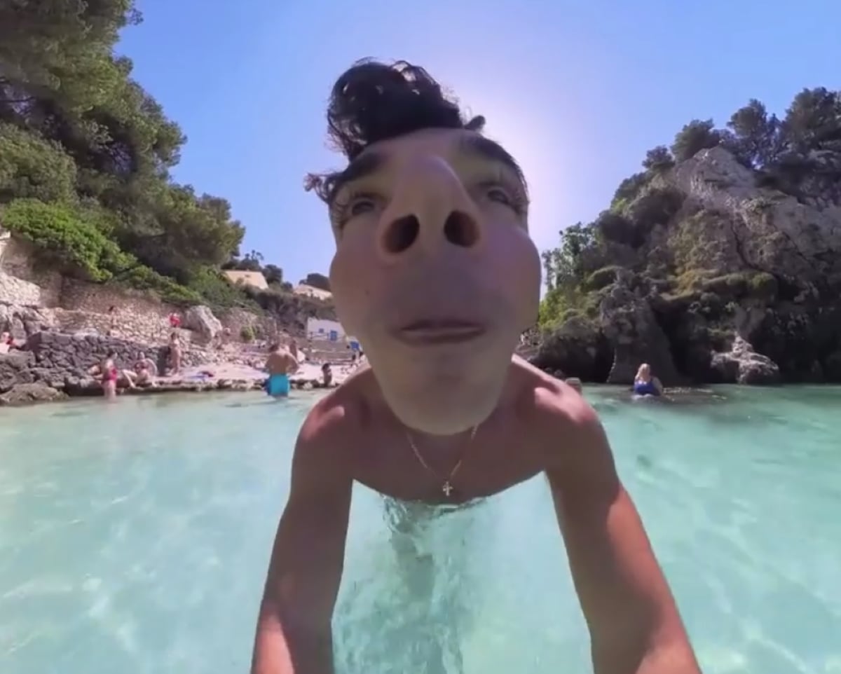 A man stands in clear water with a fisheye lens effect, making his face exaggeratedly large. Trees, rocks, and people are in the background on a sunny day.