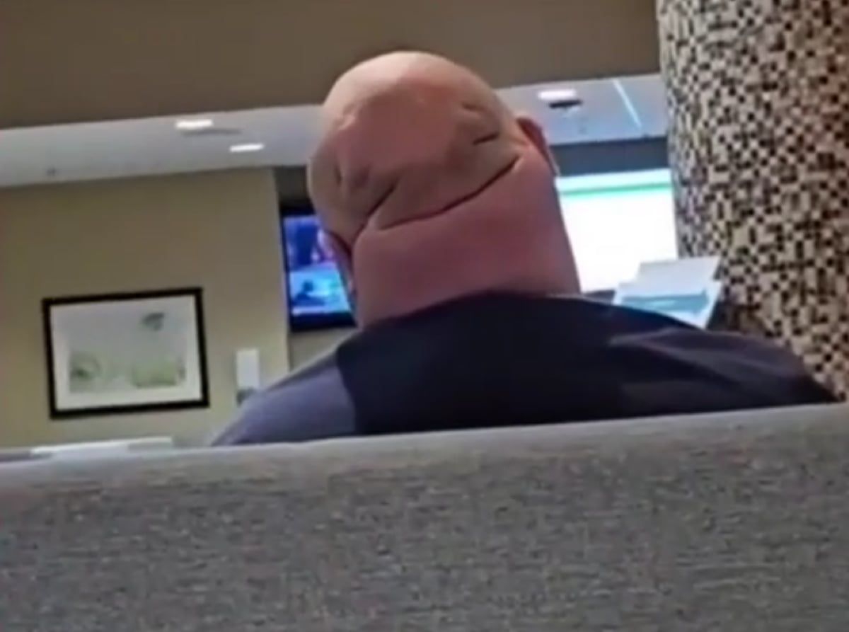A bald person is sitting on a couch, facing away. Folds on the back of their head resemble a smiling face. The background shows a TV and framed art on the wall.