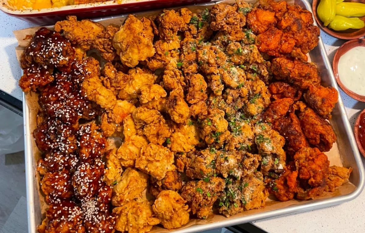 A tray filled with assorted fried chicken pieces, including saucy wings with sesame seeds, classic golden fried, herbed, and spicy red varieties, surrounded by dipping sauces.