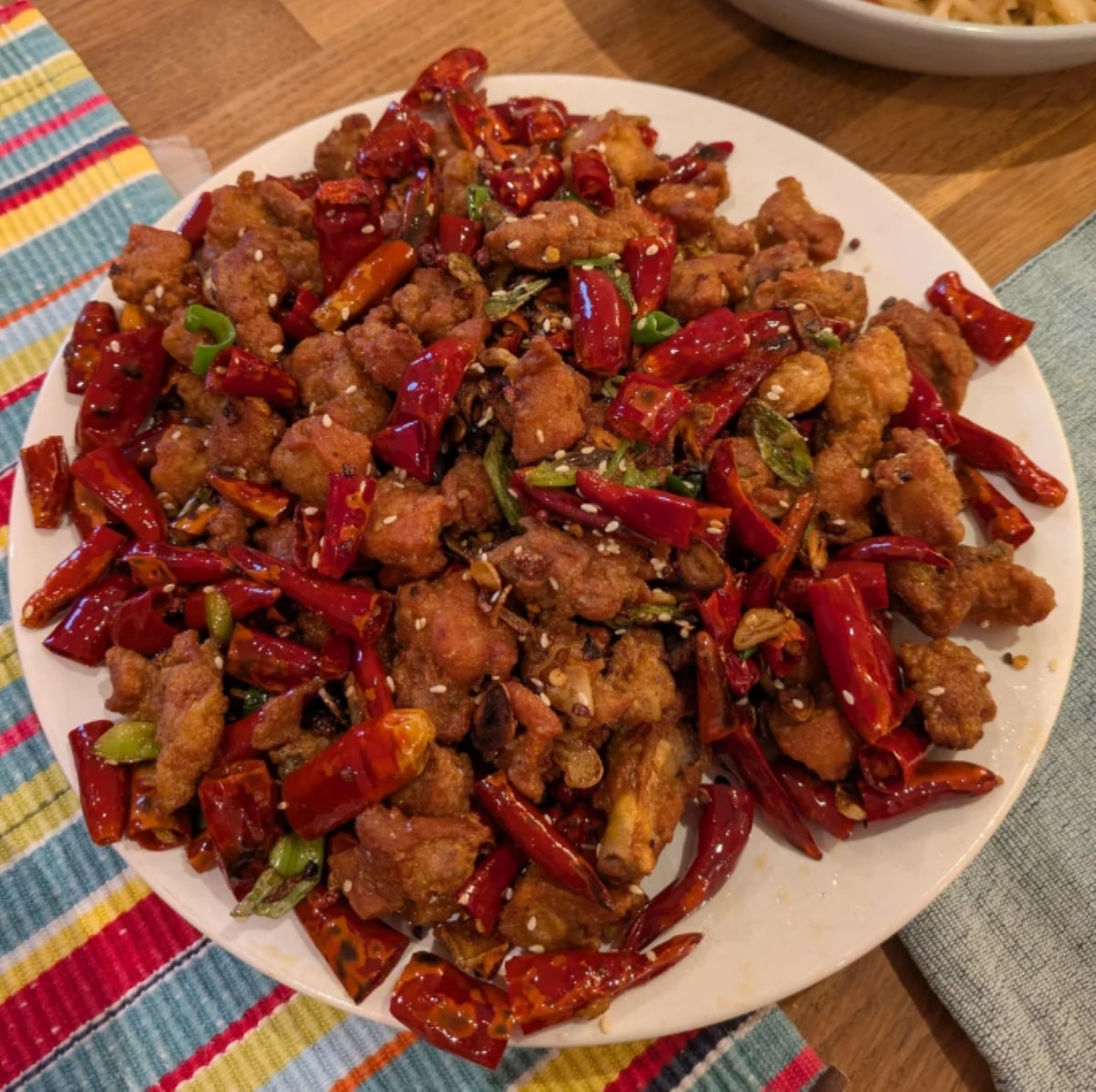 A plate of spicy fried chicken garnished with dried red chilies, green onions, and sesame seeds sits on a wooden table next to colorful striped and blue napkins.