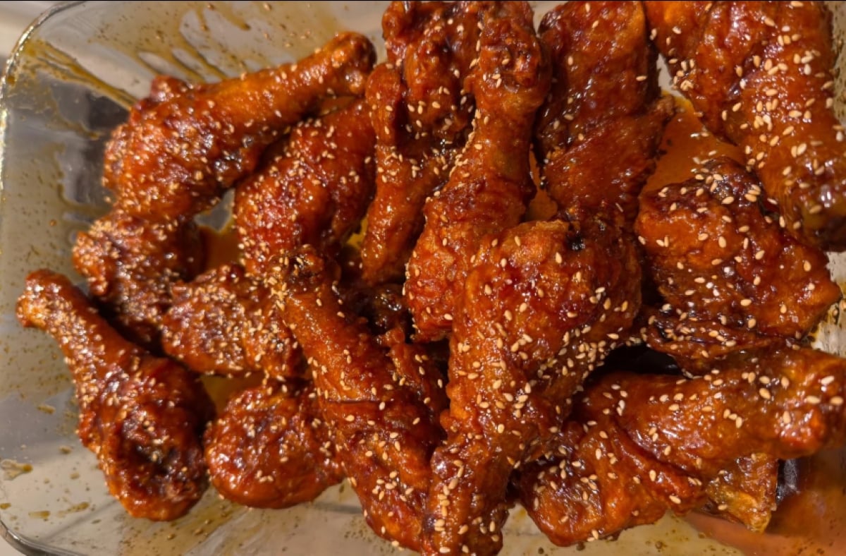 A glass tray filled with glazed chicken drumsticks coated in a sticky sauce and sprinkled with sesame seeds.