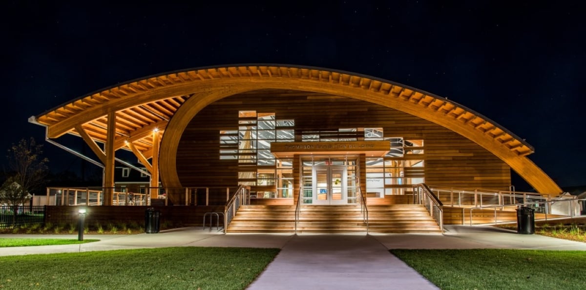 A modern building with a large, curved wooden roof is lit up at night, with glass doors and windows reflecting light. Stairs and ramps lead to the entrance, and grass lines the walkway, evoking the charm found in nostalgic travel destinations.