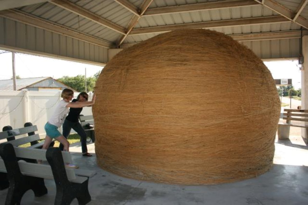 Two people are playfully pushing a giant ball of twine under a metal-roofed shelter, surrounded by benches. Sunlight filters in from the sides.