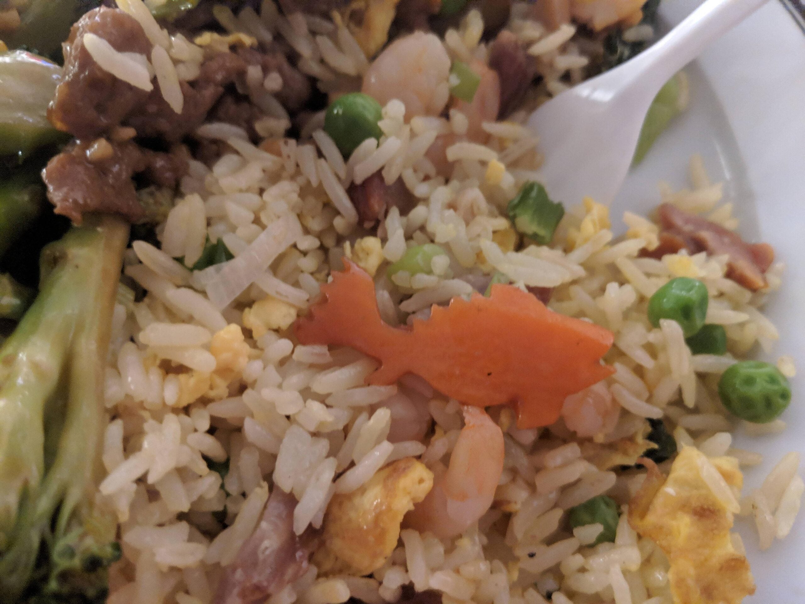 A plate of fried rice with peas, egg, shrimp, and a carrot slice cut into the shape of a fish, with broccoli and a plastic fork visible.