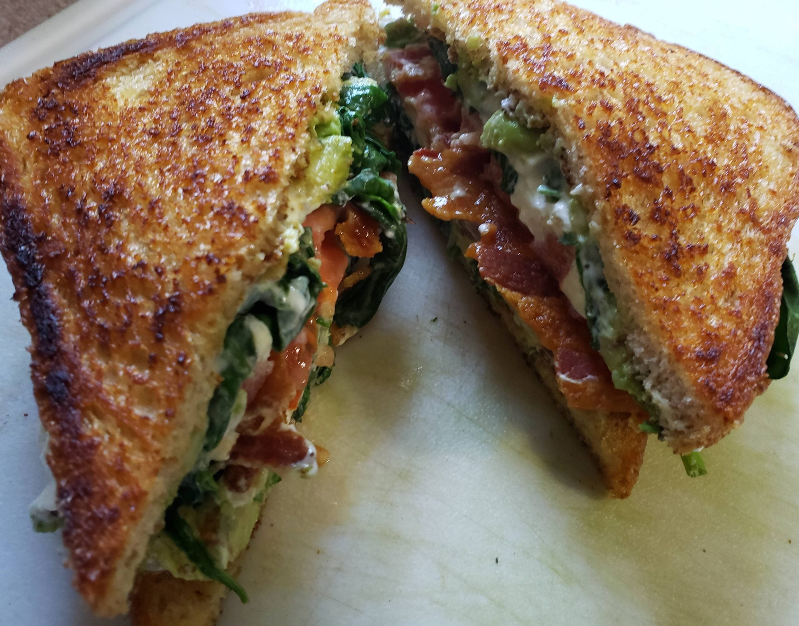 A grilled sandwich cut in half, filled with crispy bacon, lettuce, tomato, and creamy sauce, is displayed on a cutting board.