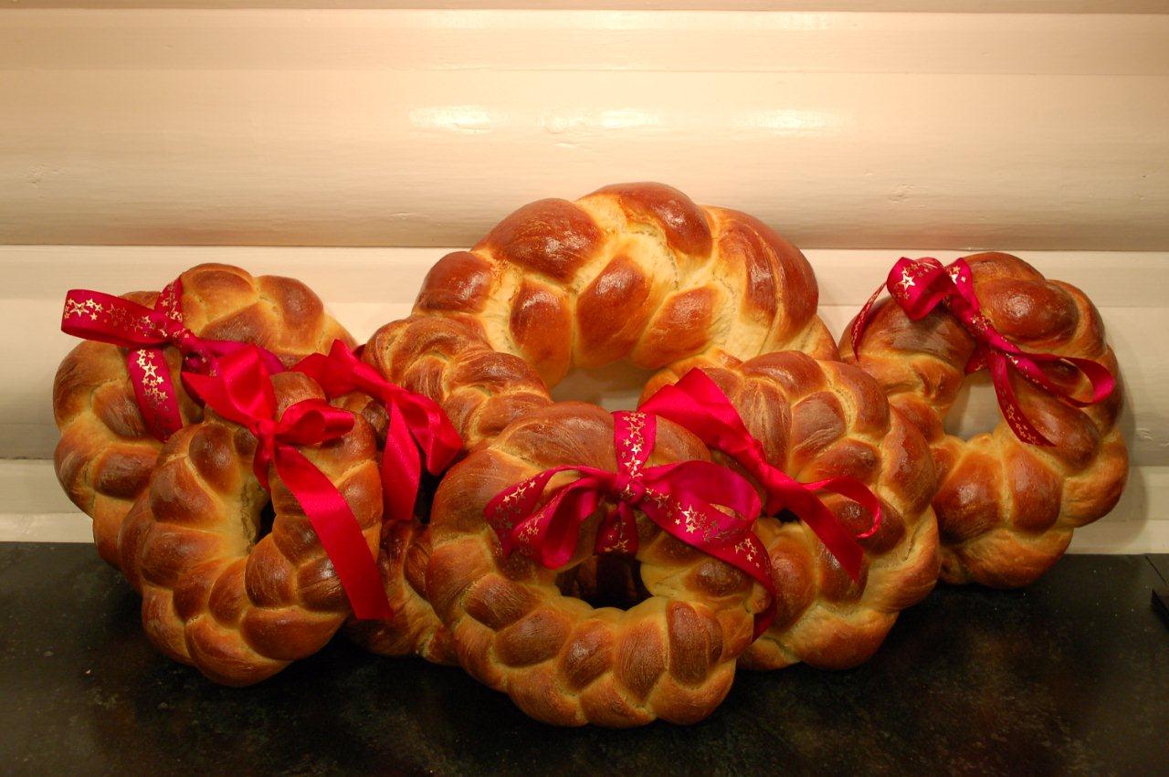 Five braided bread wreaths, each decorated with a red ribbon, are arranged on a dark surface in front of a light-colored wooden background.