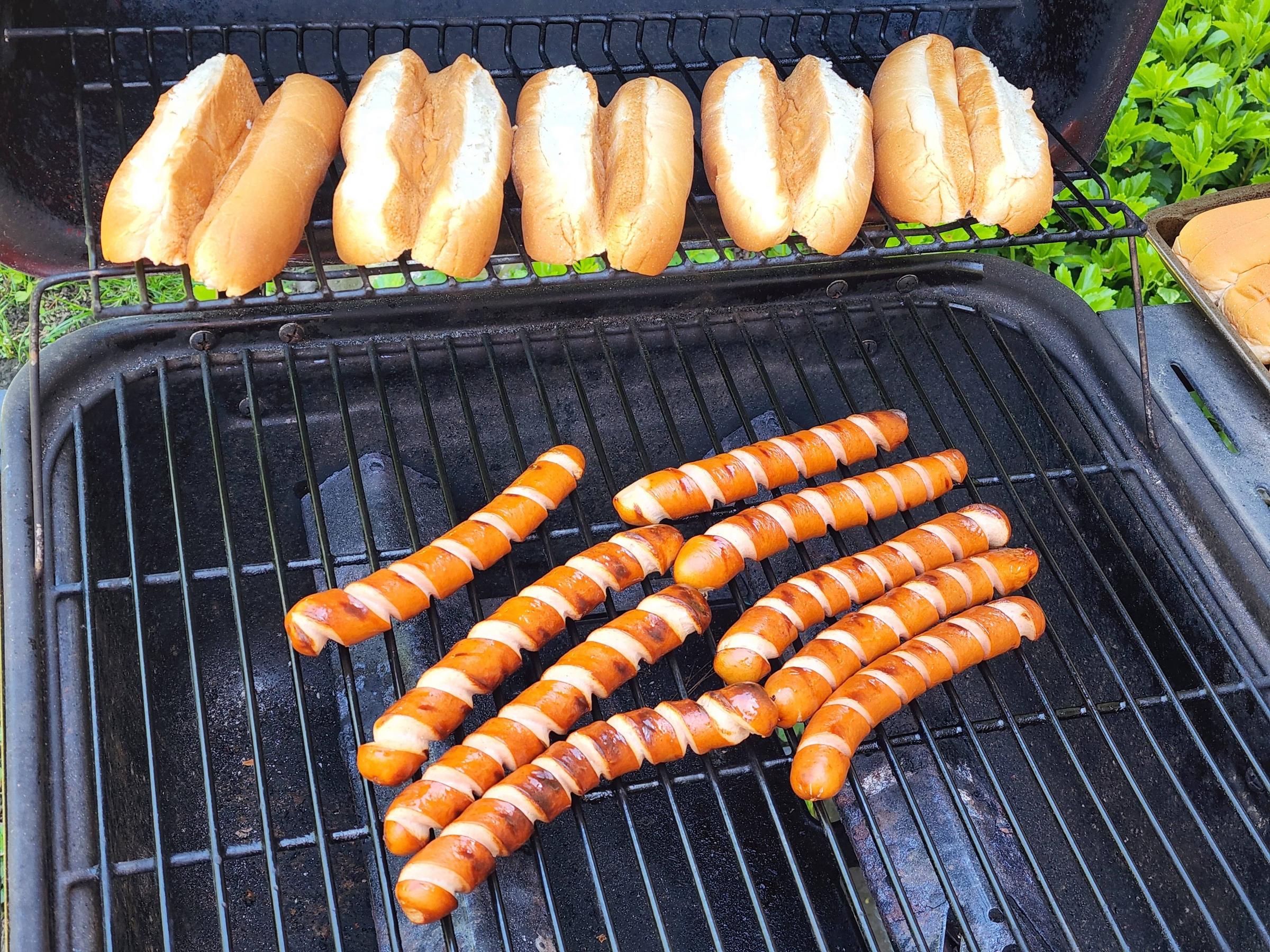 Hot dog buns on the top grill rack and several grilled hot dogs with grill marks on the lower rack of an outdoor barbecue grill.