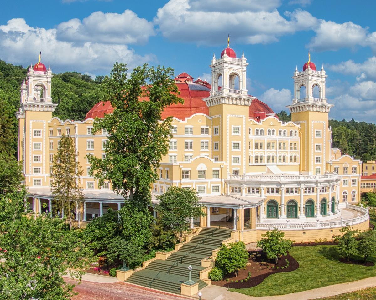 A grand yellow building with red domes, white trim, and tall towers—reminiscent of historic hotels—stands surrounded by trees and greenery under a blue sky with fluffy clouds.