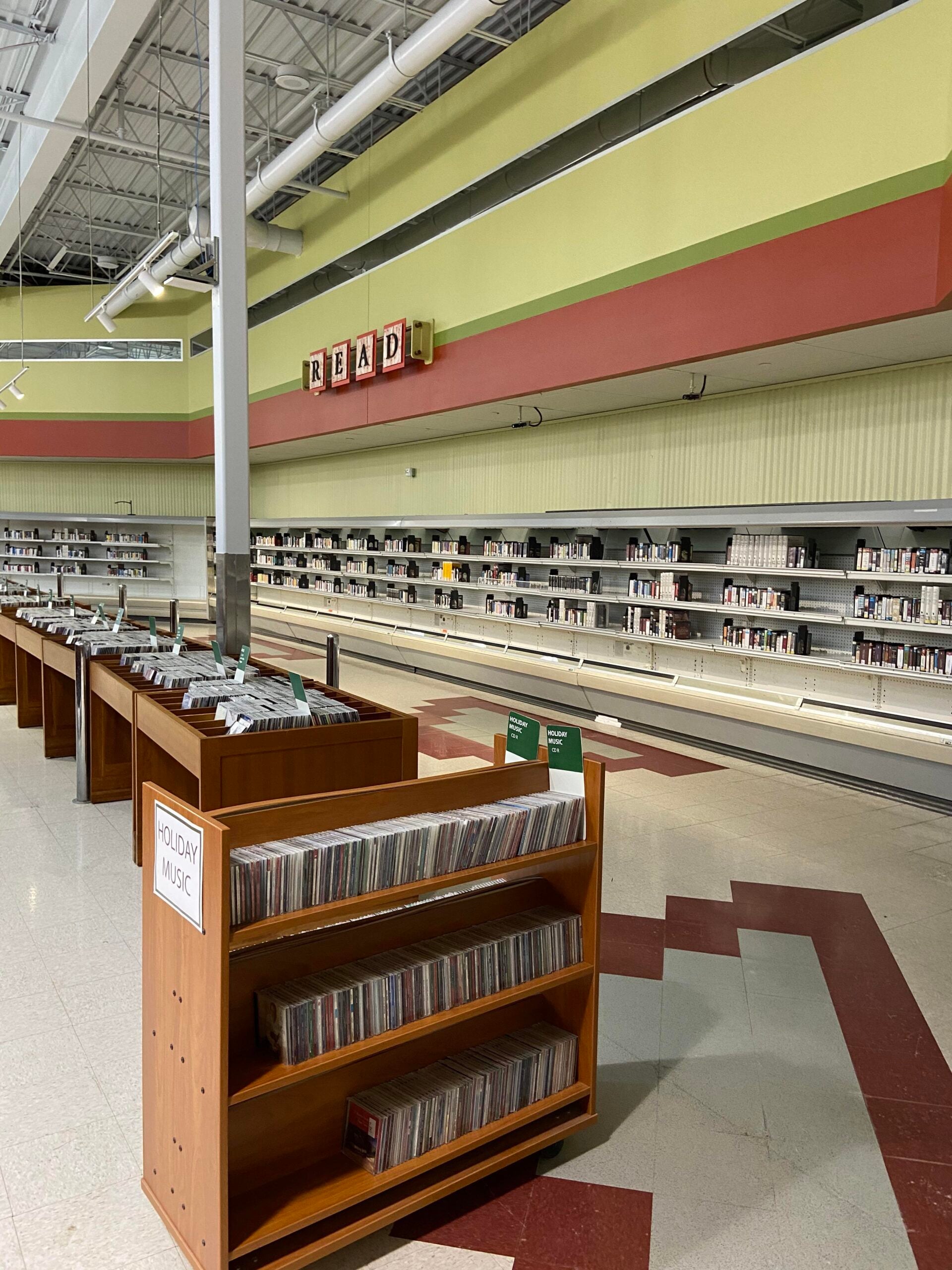 A library with shelves of books and CDs set up in an old grocery store. A sign spelling “READ” hangs above empty refrigerated cases now used as bookshelves. The floor has red and white tiles.