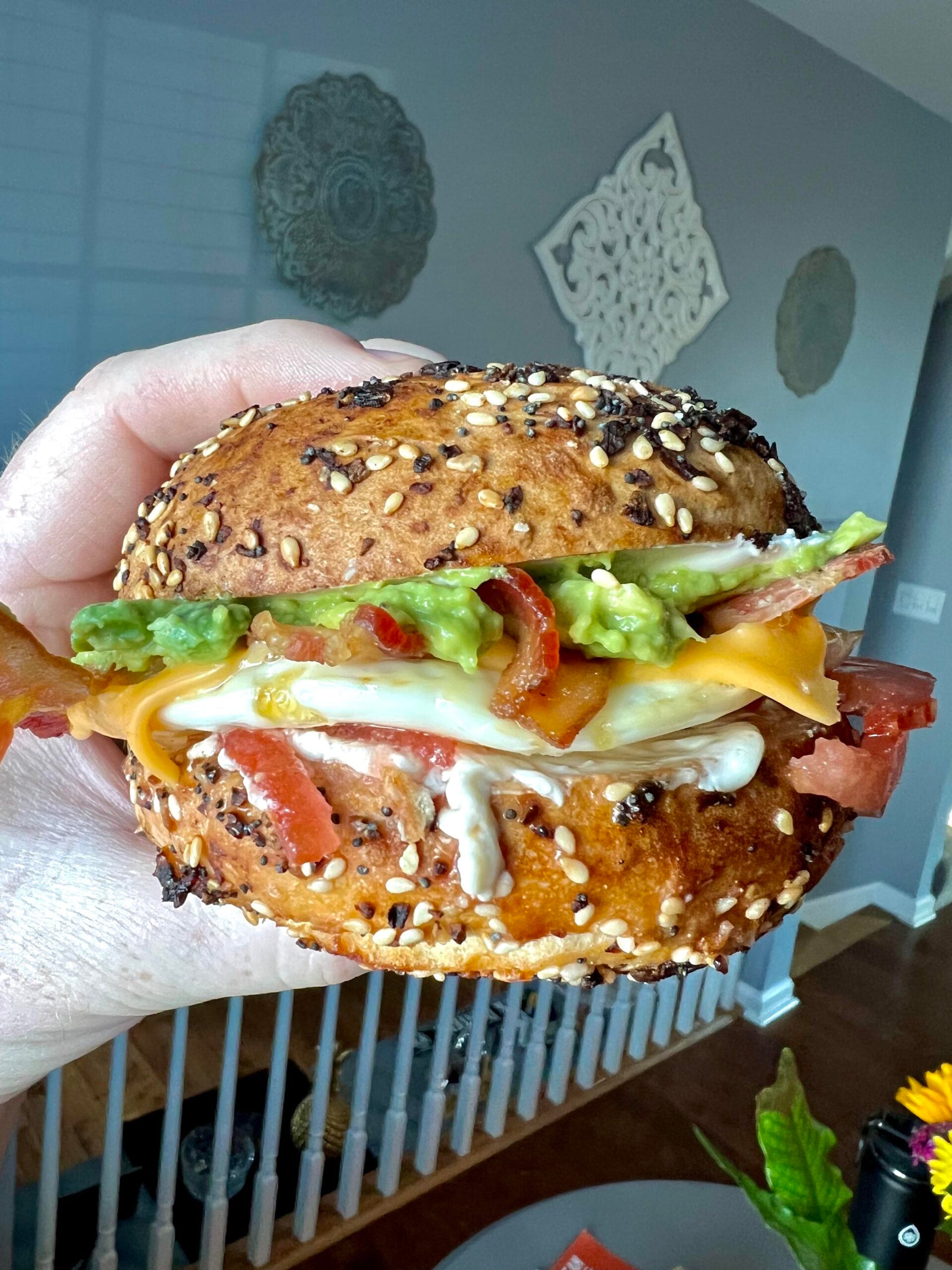 A hand holds a sandwich with a seeded bun, cheese, bacon, mayonnaise, tomato, turkey, and mashed avocado. A home interior with gray walls and decorative art is visible in the background.
