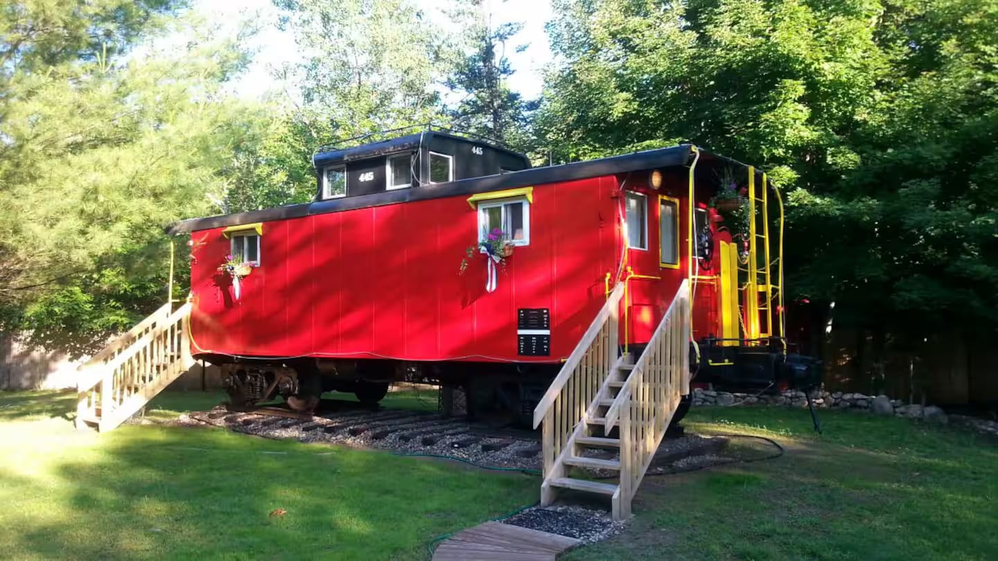 A bright red caboose train car converted into a tiny home, with wooden steps leading to both doors, sits on tracks surrounded by green grass and trees.