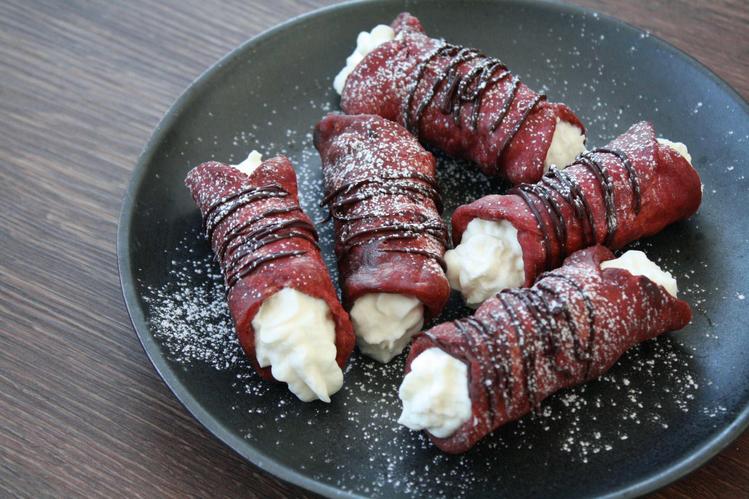 Five homemade cannolis, made with red velvet shells and filled with cream, are drizzled with chocolate and arranged on a black plate, lightly dusted with powdered sugar, on a wooden table.