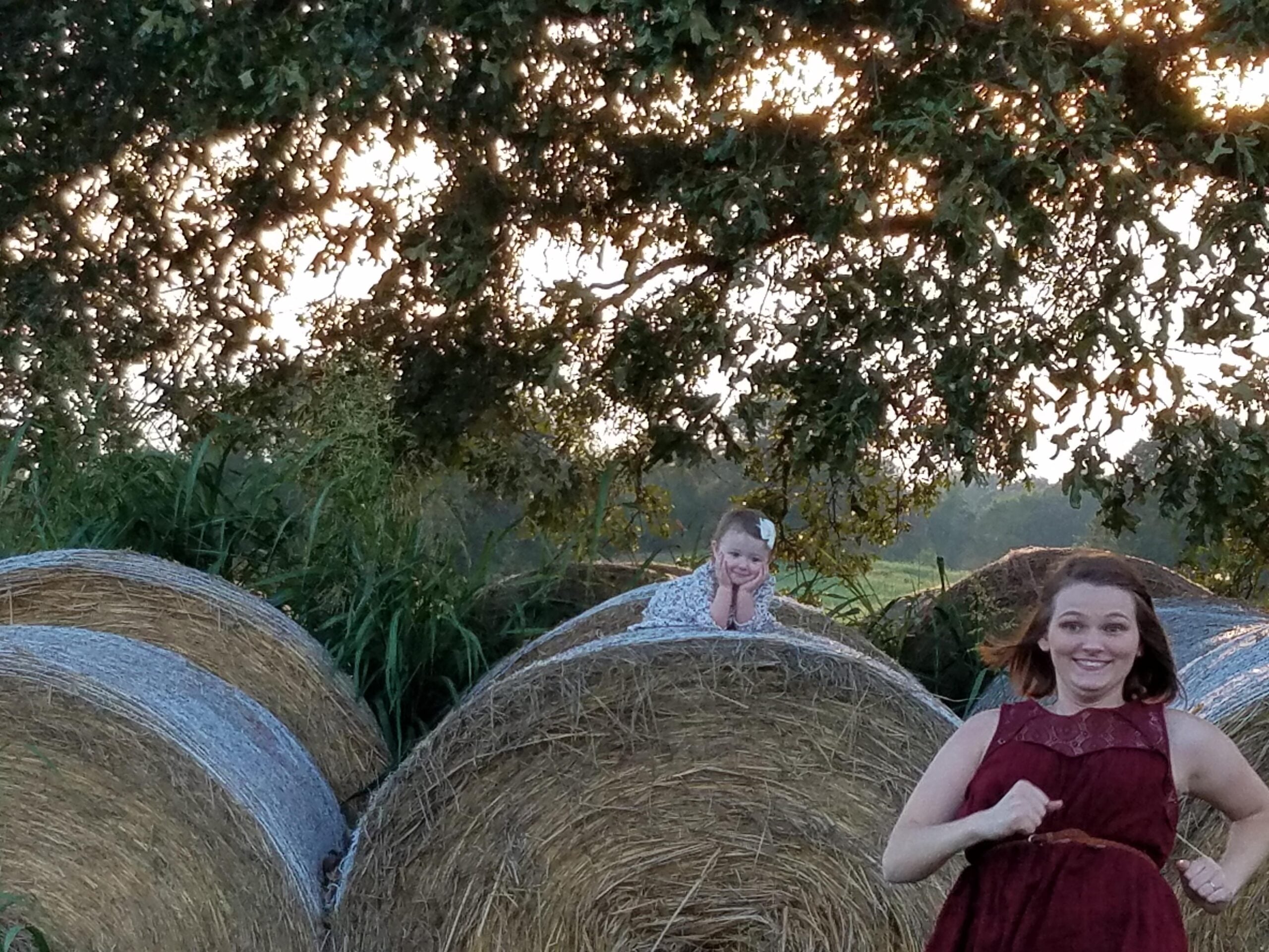 Two girls are pictured outdoors; one girl in a red dress runs and smiles in the foreground, while another girl sits atop a large round hay bale under a tree at sunset.