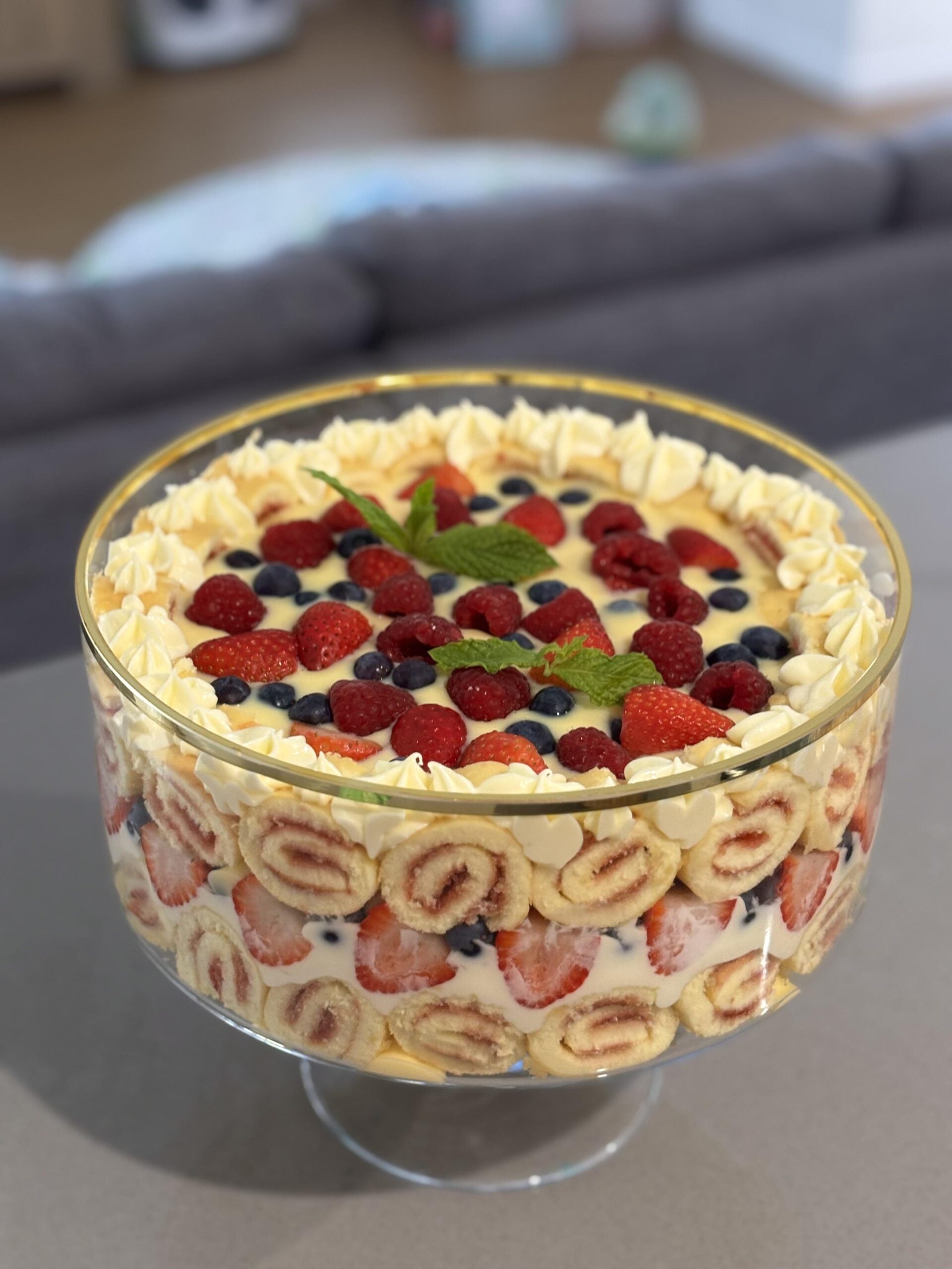 A glass trifle bowl filled with layers of cream, sliced jam roll, strawberries, blueberries, and raspberries, topped with piped cream, berries, and a sprig of mint—this showstopping dessert is the perfect centerpiece for your homemade Christmas treats.