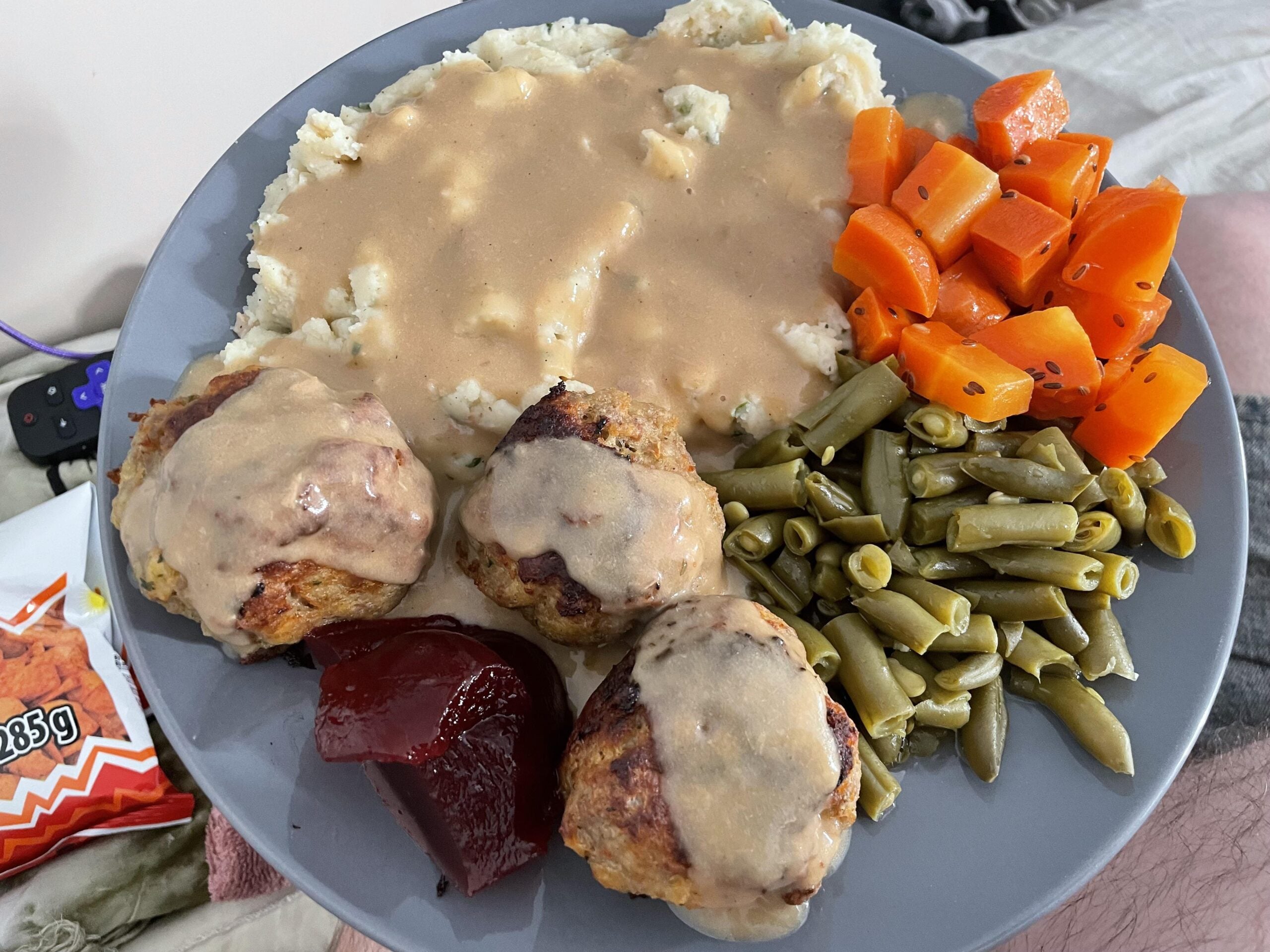 A plate with mashed potatoes and gravy, glazed carrots, green beans, three meatballs with gravy, and a slice of cranberry sauce. A TV remote and snack wrapper are in the background.