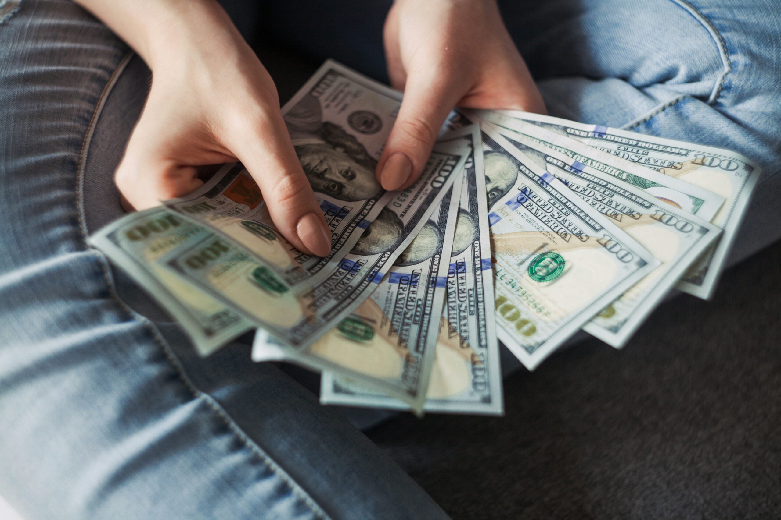 A person in blue jeans is holding and fanning out several $100 US dollar bills while sitting cross-legged on the floor, perhaps contemplating the challenges of life in 2026 problems and financial decisions that come with the changing times.