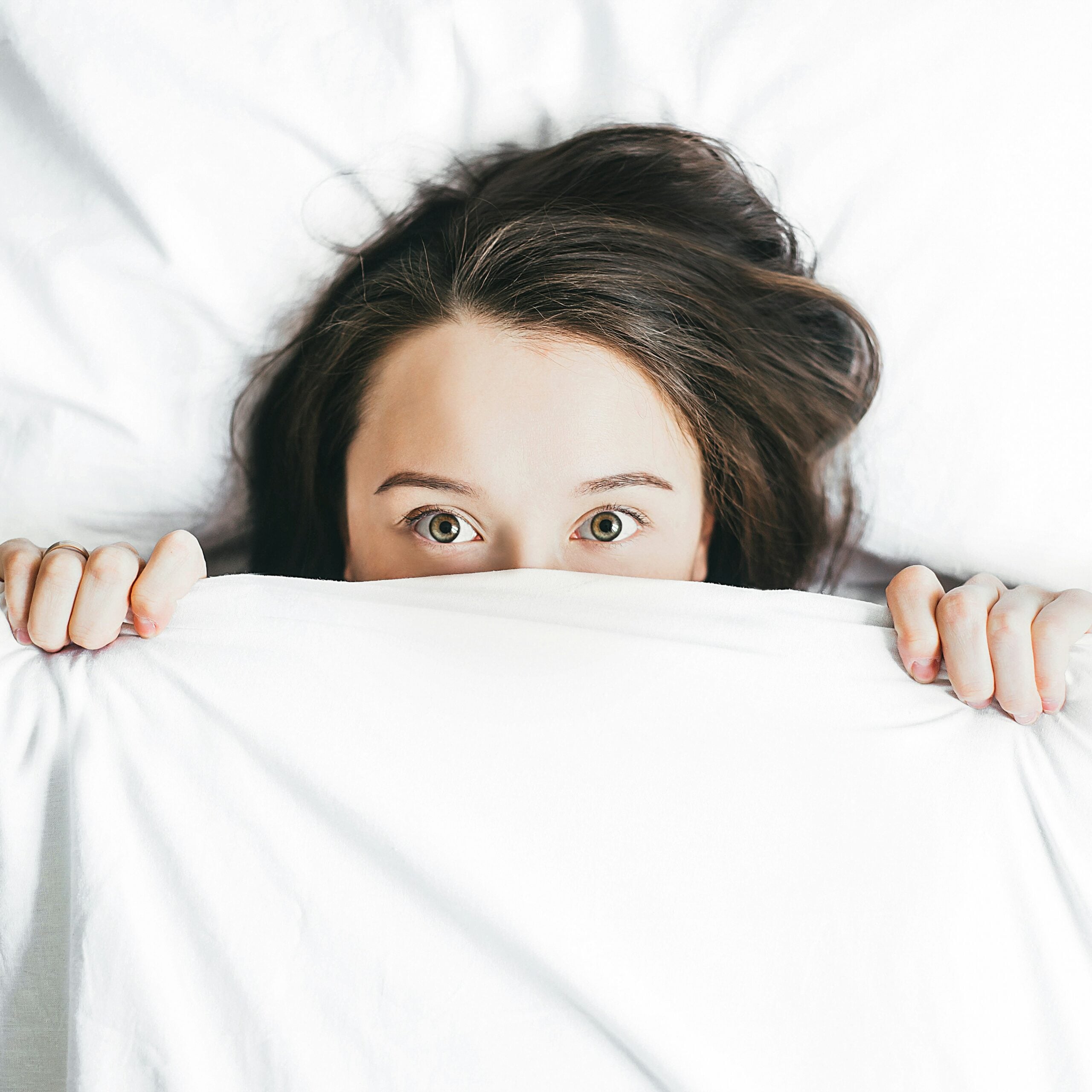 A person with long dark hair lies in bed, peeking out from under a white blanket, with only their eyes and part of their face visible, perhaps quietly contemplating life in 2026 problems as they seek comfort in the safety of their covers.