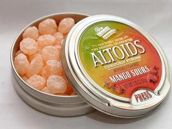 An open tin of Altoids Curiously Strong Mango Sours candies, showing several orange, round candies inside the metal container with the colorful lid partially covering them.
