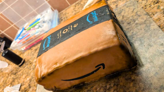 A realistic decorated cake designed to look like an Amazon Prime package sits on a granite countertop, complete with the Amazon logo, shipping tape, and brown box details.
