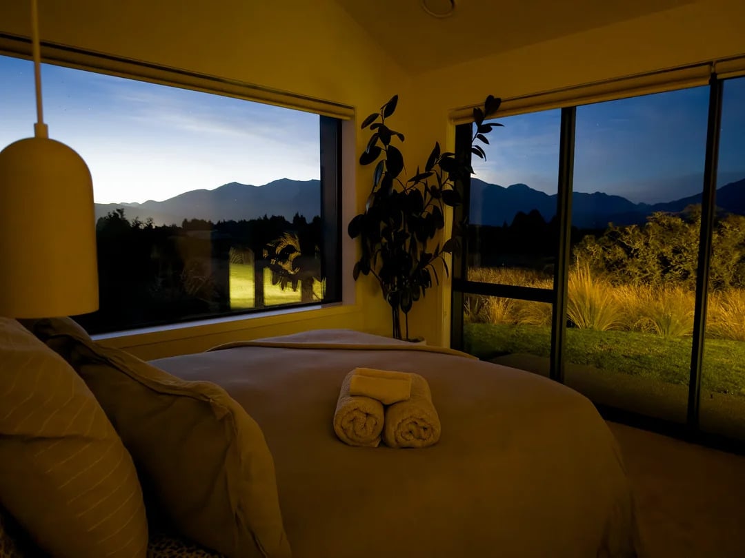 A cozy bedroom at dusk with large windows showing mountain views, a bed with beige linens and two rolled towels, and a potted plant by the window.