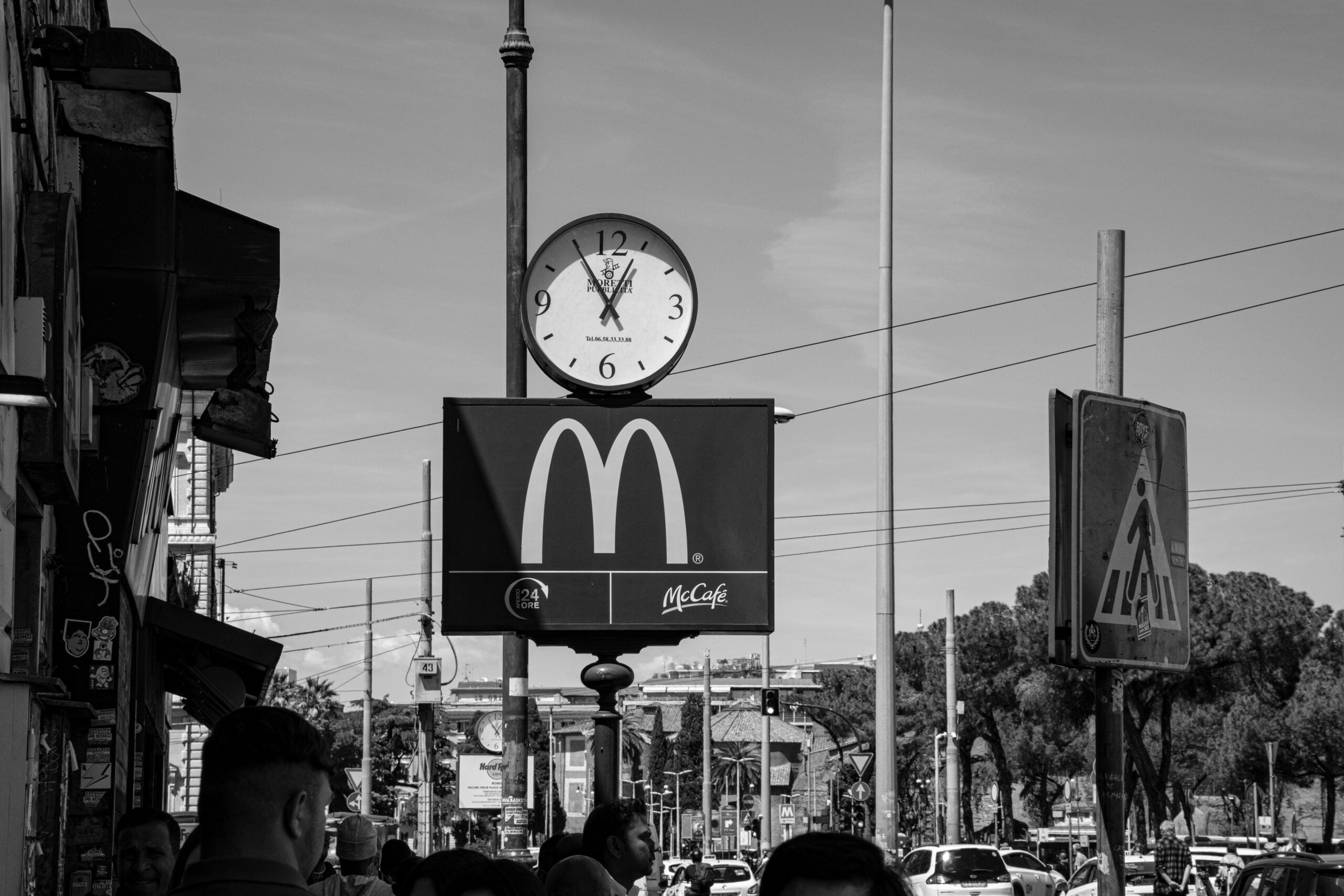 Black and white photo of a McDonald’s sign with a clock above it showing 11:57. People and cars are visible in the foreground, with street signs and trees in the background.