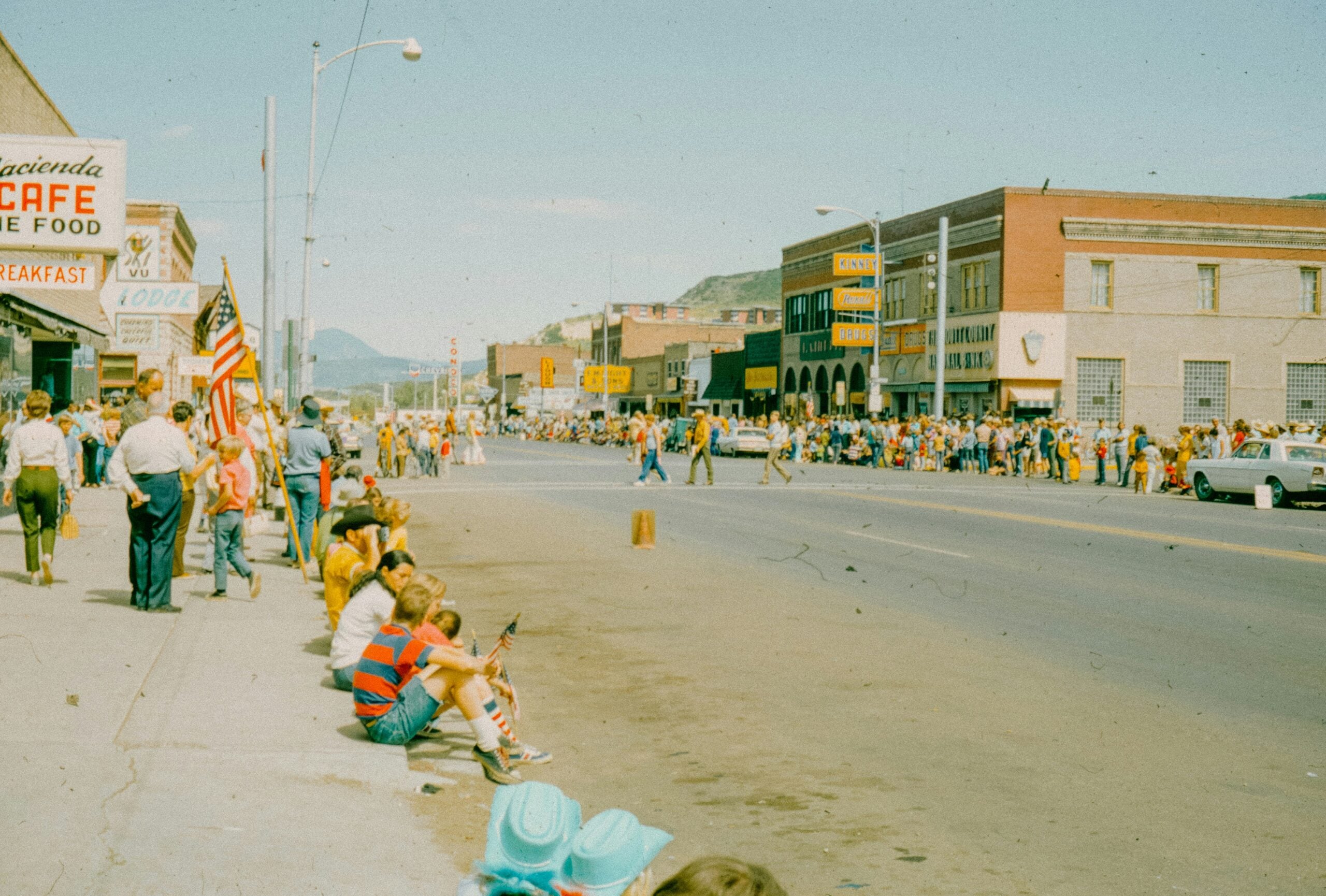 A crowd lines the street, some seated on the sidewalk, watching a parade. Flags and signs are visible; buildings and mountains are in the background. The sky is clear and sunny.