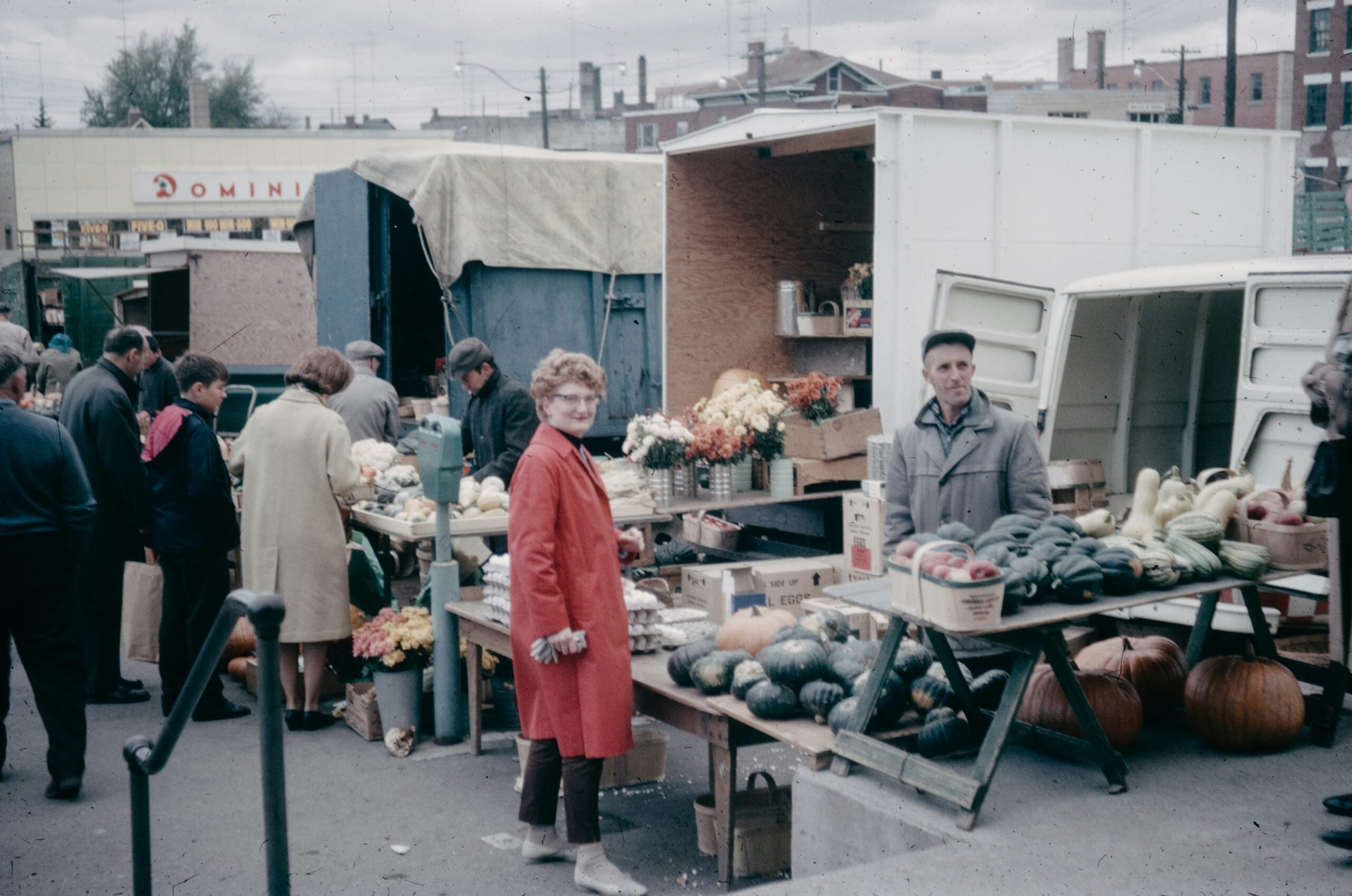 People shop at an outdoor market with fruit, vegetables, and flowers on tables. A woman in a red coat smiles at the camera, while vendors stand by their stalls. Trucks are parked behind the market stands.