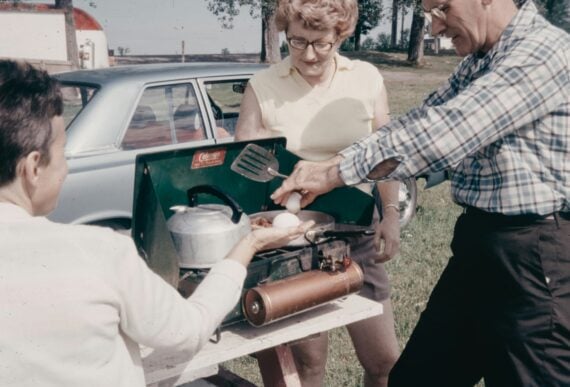 Three adults cook eggs outdoors on a portable stove beside a car. One person holds a spatula, another cracks an egg, and a third holds a pan. Trees and grass are visible in the background.