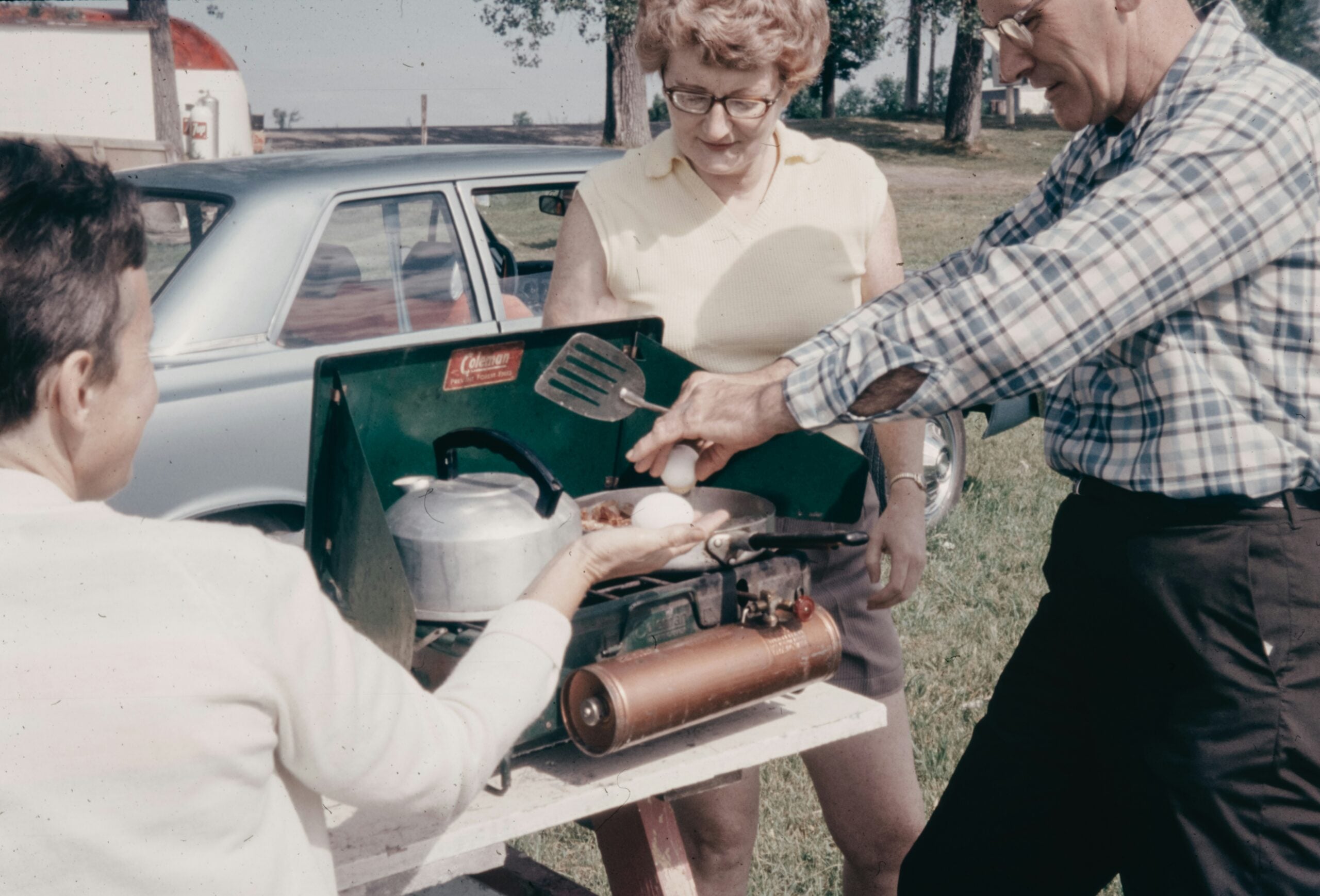 Three adults cook eggs outdoors on a portable stove beside a car. One person holds a spatula, another cracks an egg, and a third holds a pan. Trees and grass are visible in the background.
