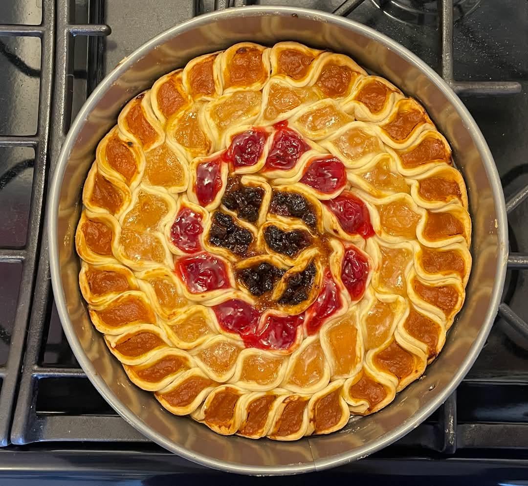 A round pie with a decorative, wavy crust pattern and filling in multiple colors—red, amber, and dark purple—sits in a metal pan on a stovetop.