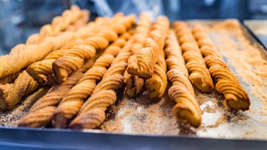 A close-up of several twisted churros coated in cinnamon sugar, arranged in rows on a metal tray.