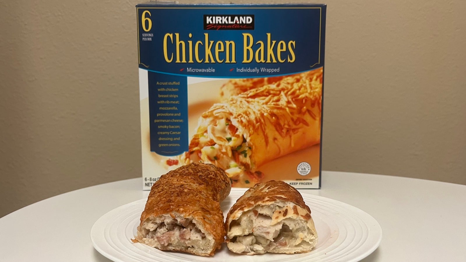 A plate with a sliced open chicken bake sits in front of a Kirkland Signature Chicken Bakes box showing an image of the product and text highlighting it is microwavable and individually wrapped.