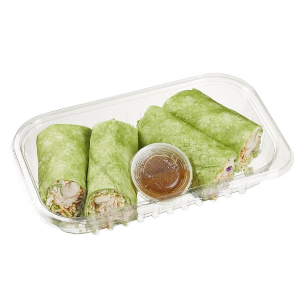 A clear plastic container with four green spinach wraps filled with vegetables, sliced in halves, and a small cup of brown dipping sauce in the center.