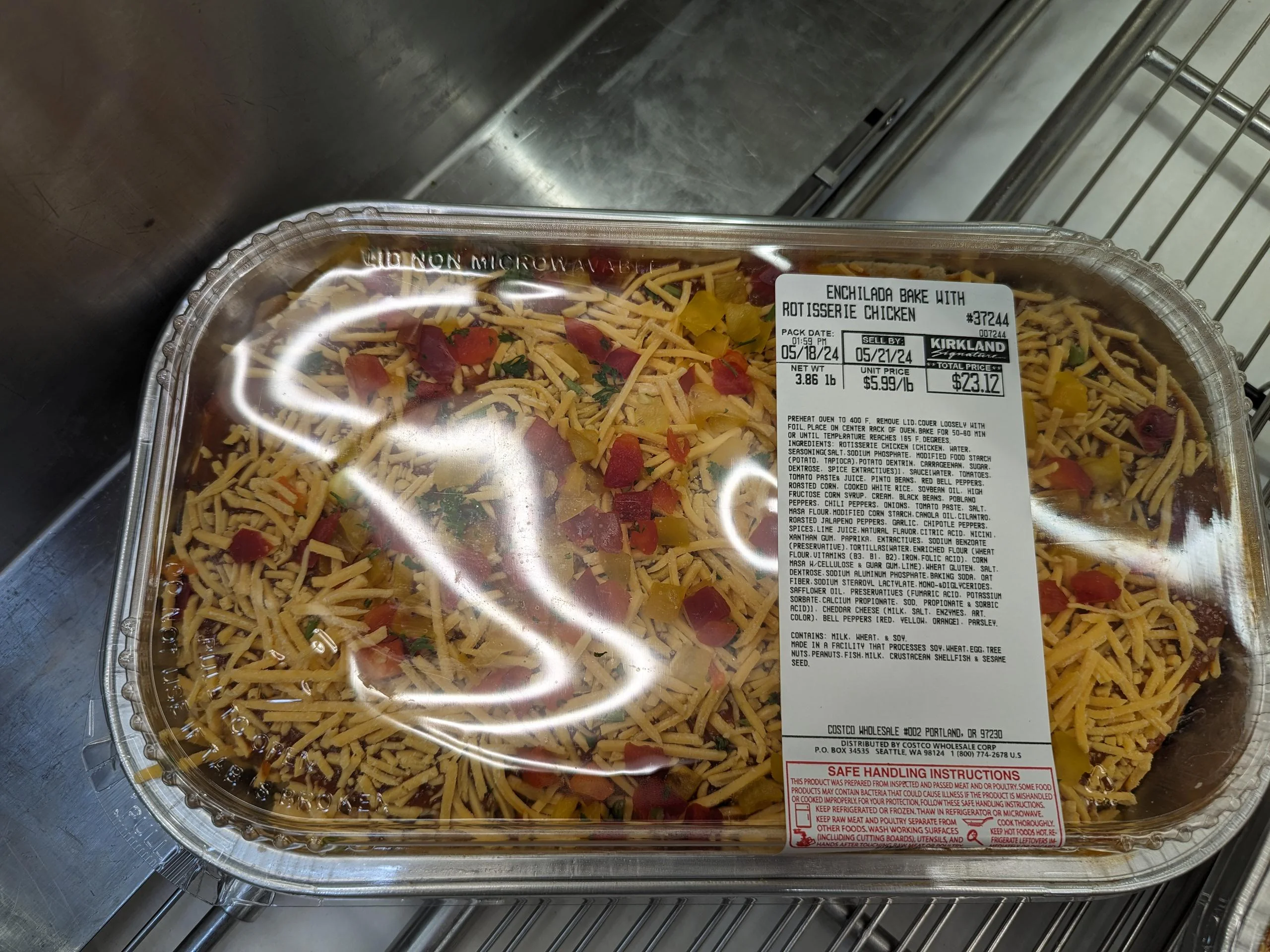 A packaged enchilada bake with rotisserie chicken sits on a metal shelf. The clear lid shows shredded cheese and diced vegetables on top. A white label with ingredients and pricing is attached.