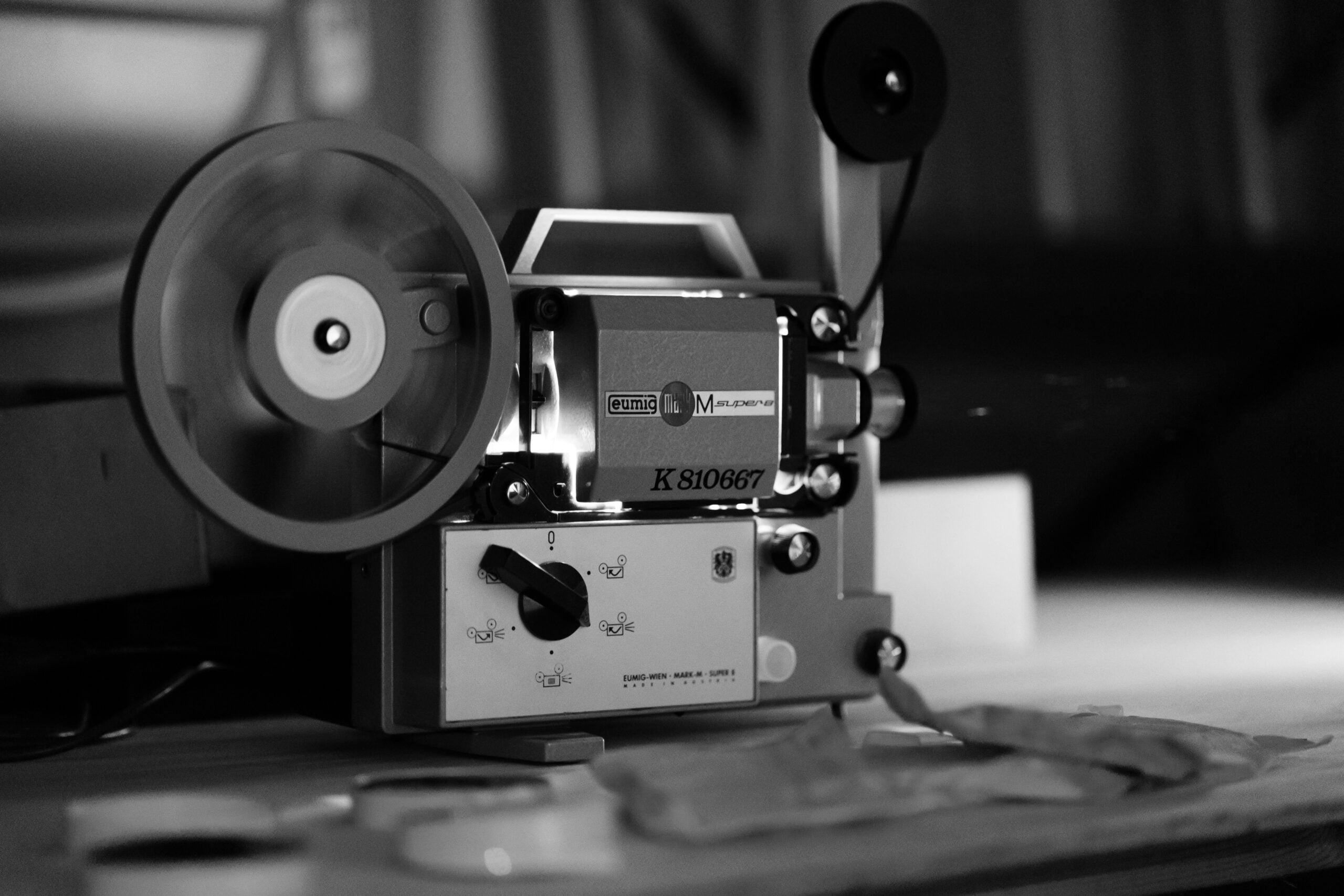 A black and white photo shows a vintage film projector in motion on a table, with reels spinning and film stretched across the machine. Film canisters and strips are scattered nearby.