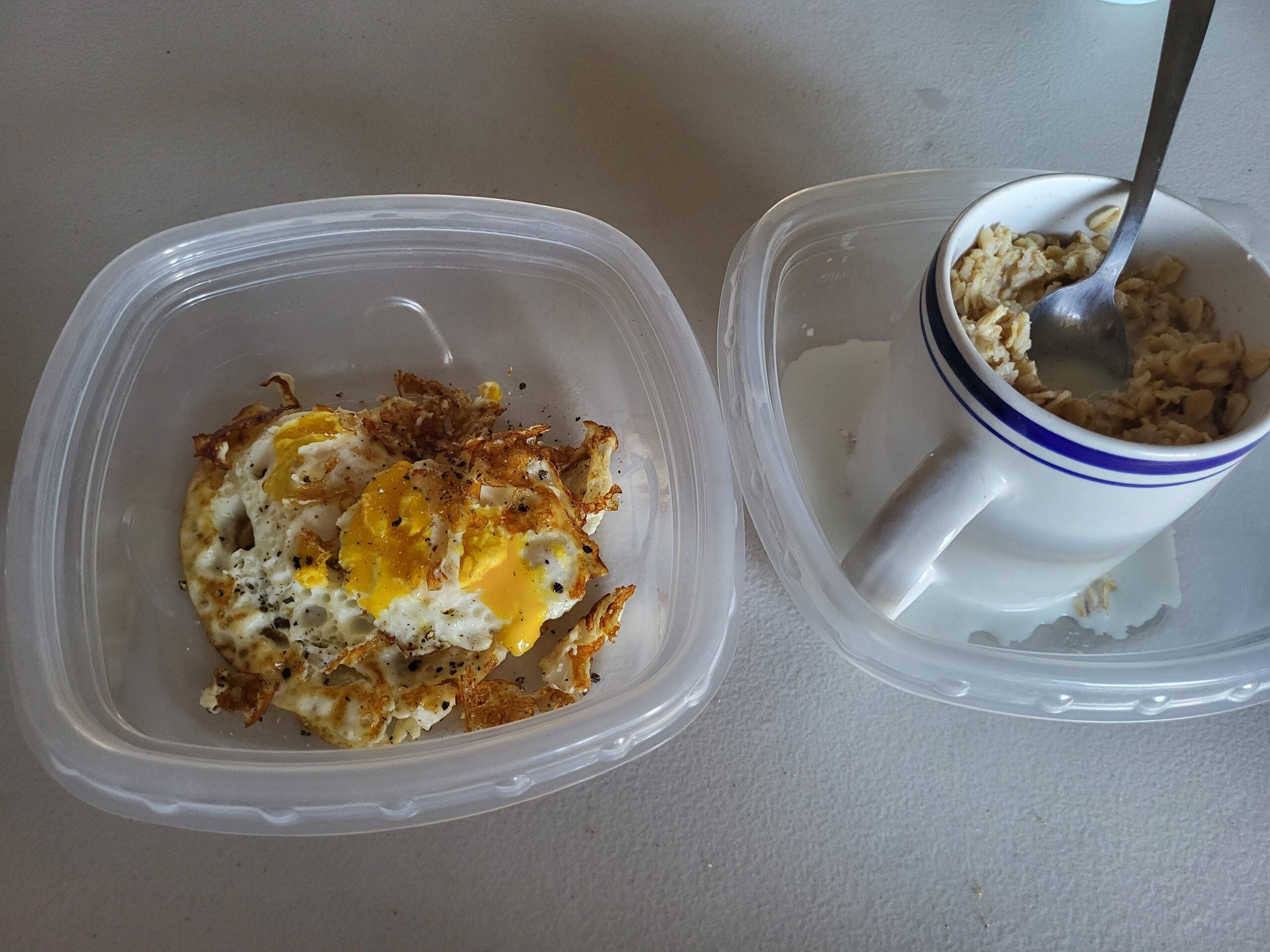 Two plastic containers: one holds two fried eggs with crispy edges, the other has a ceramic mug filled with oatmeal and a spoon. Both are set on a white surface.