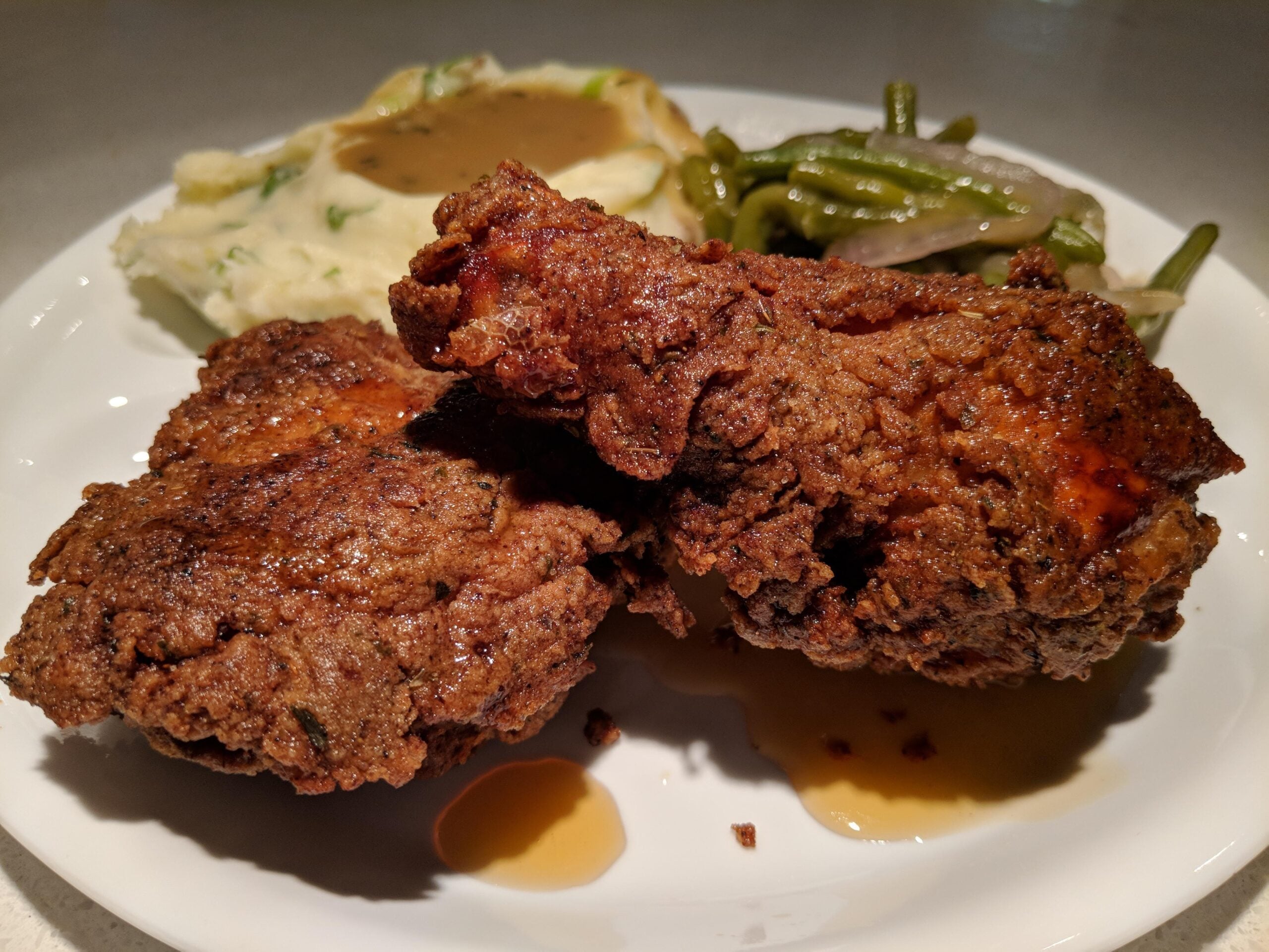 Two pieces of fried chicken on a white plate with a side of mashed potatoes topped with gravy and green beans.