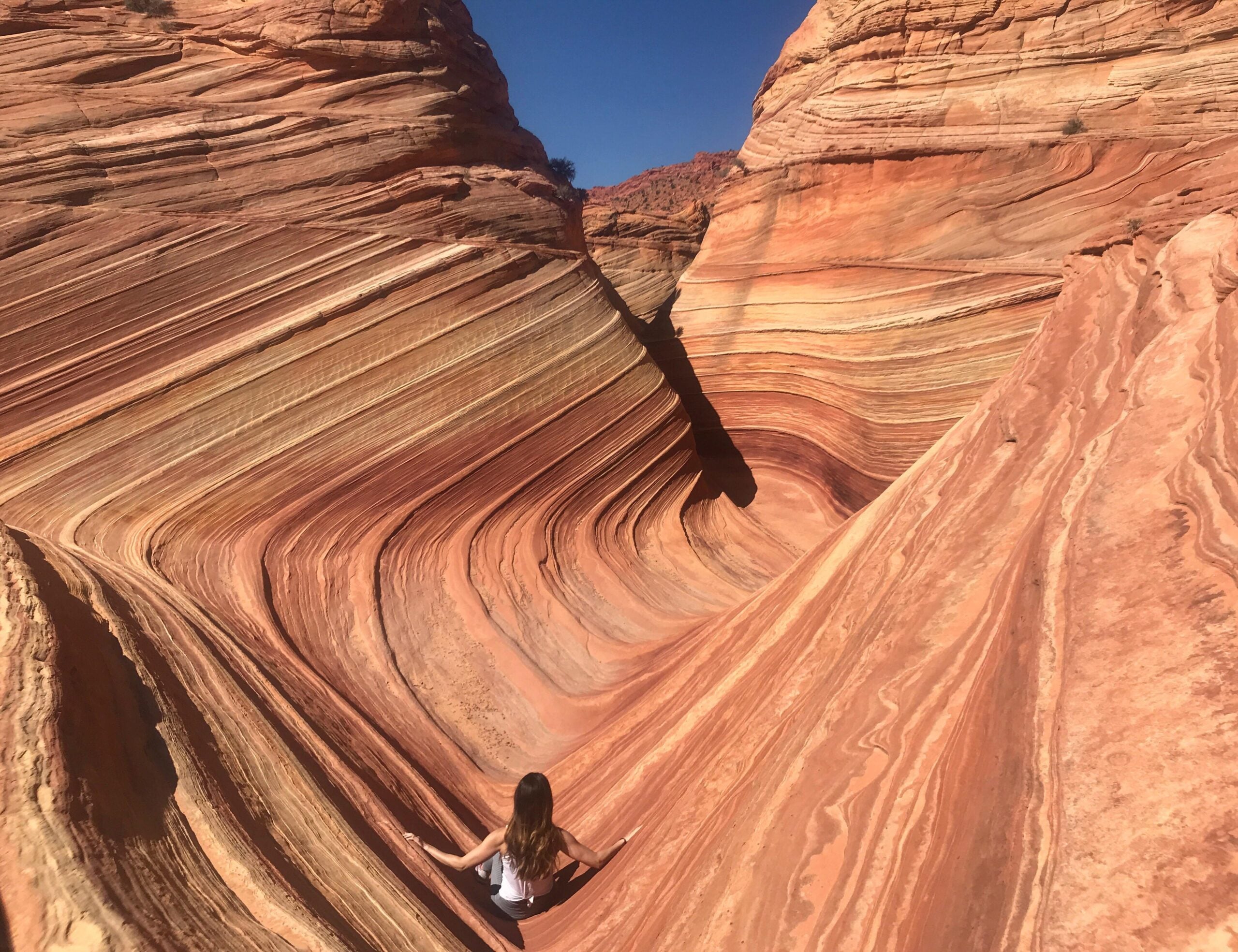 A person sits facing layered, swirling red and orange sandstone formations in a desert landscape under a clear blue sky.