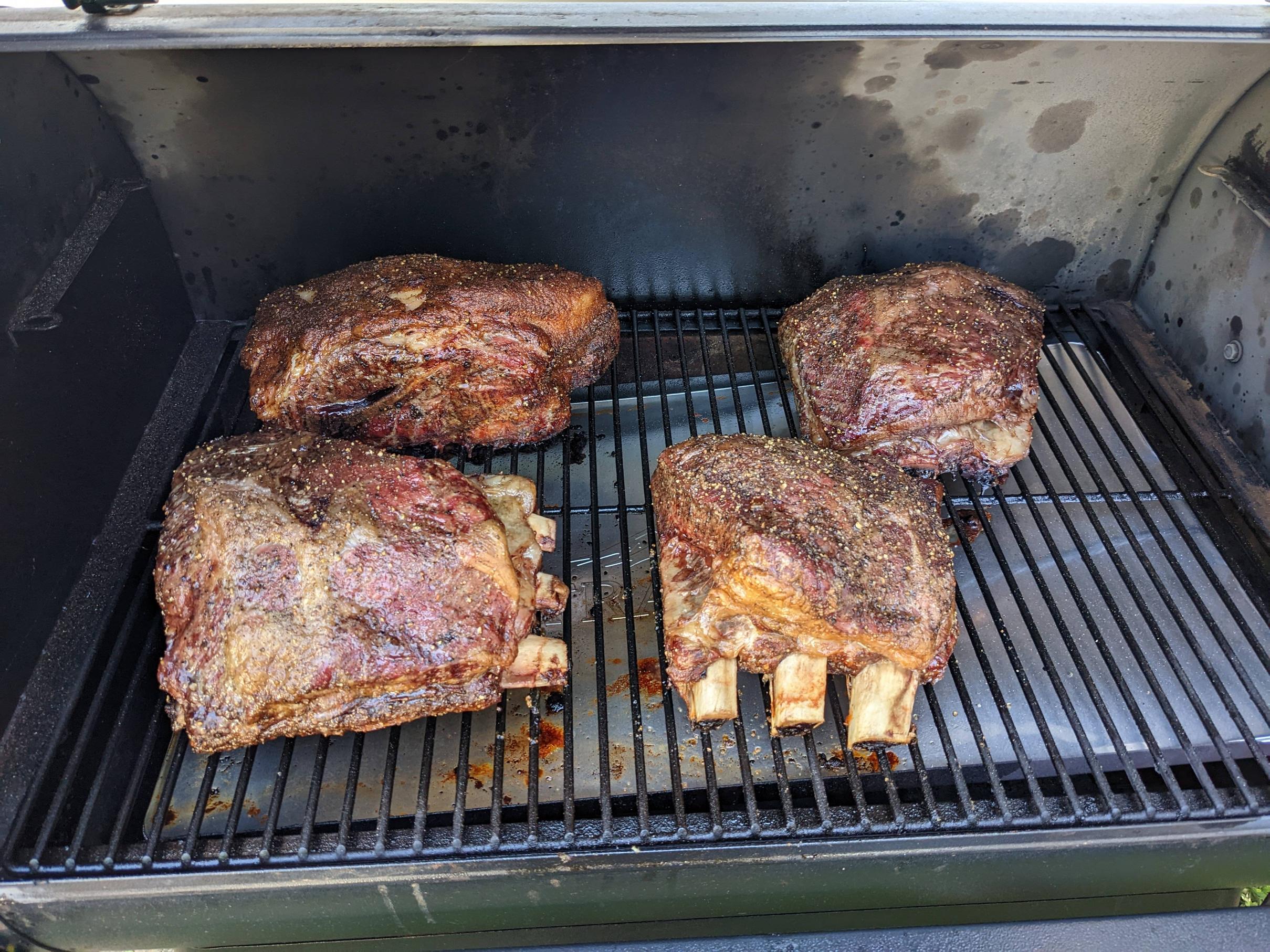 Four large, seasoned beef ribs are being smoked on the grates inside a barbecue smoker. The meat has a dark, crisp exterior and is positioned evenly on the smoker rack.