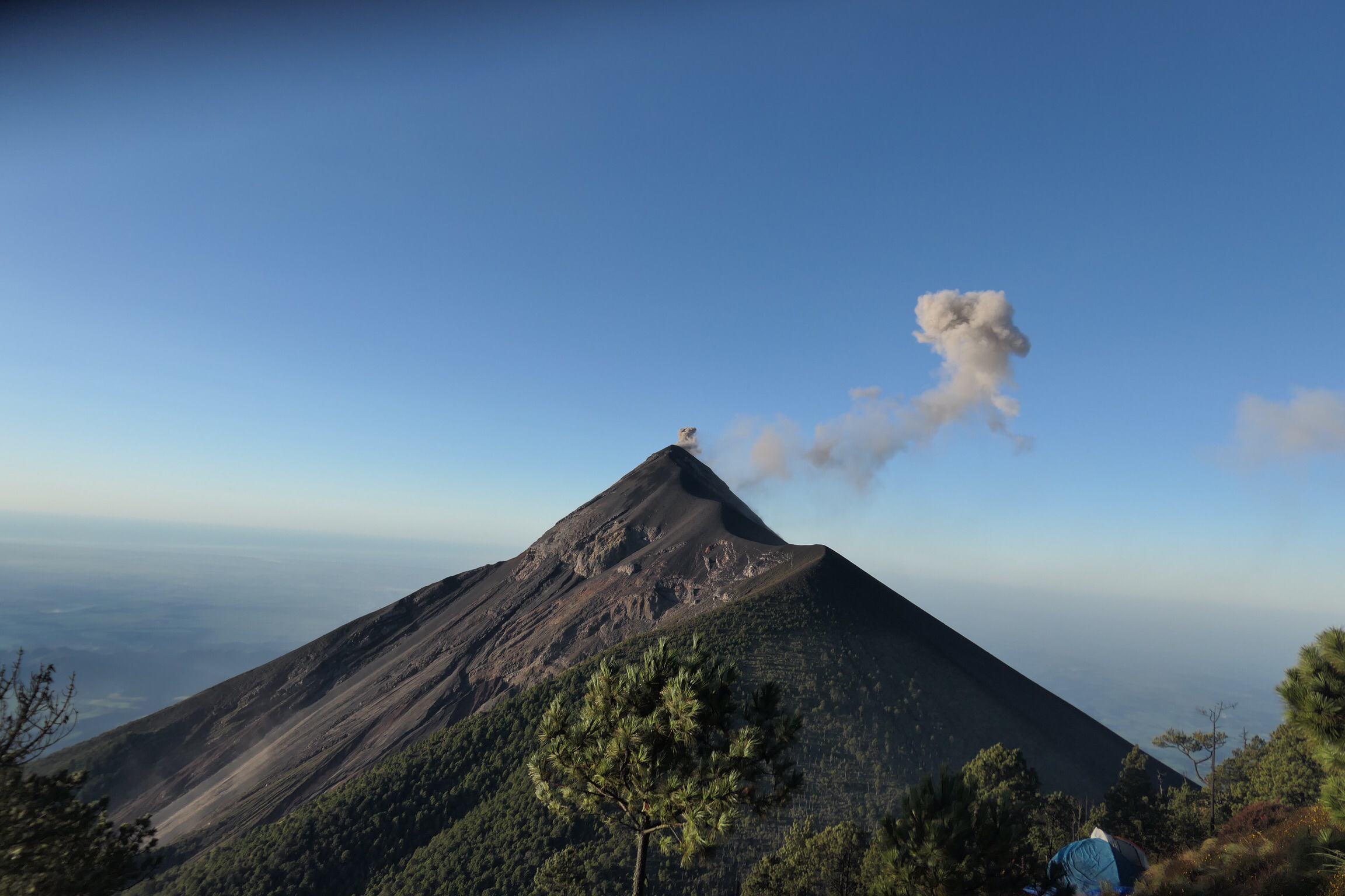 A conical volcano emits a plume of gray smoke under a clear blue sky, with green trees in the foreground and sunlight highlighting the mountain slopes.