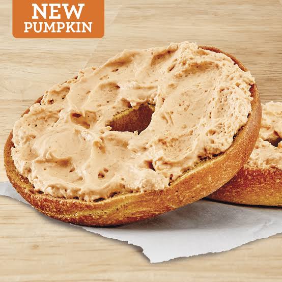A pumpkin bagel topped with pumpkin-flavored cream cheese sits on parchment paper, with a “NEW PUMPKIN” label in the top left corner.